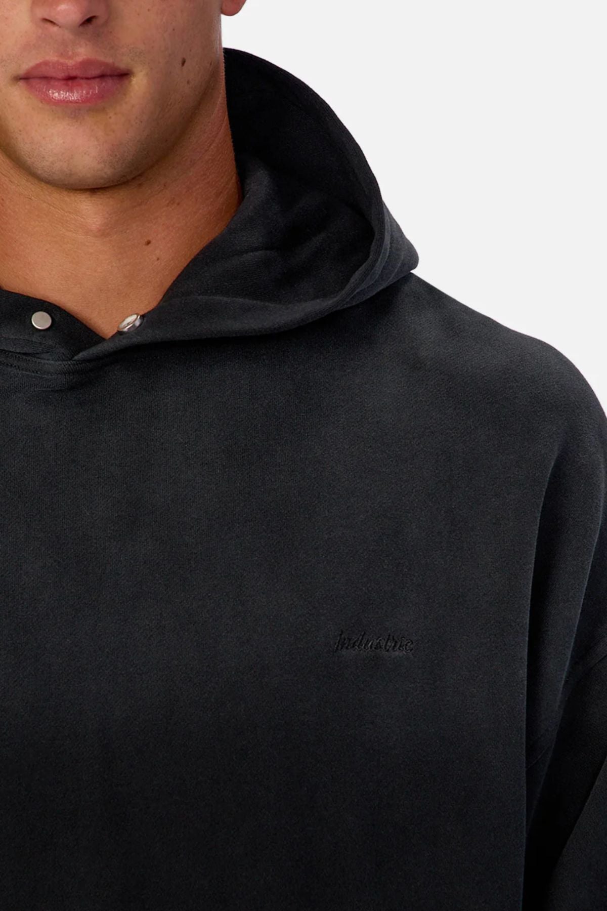 A close-up shows a person wearing the Industrie The Sandblast Hoodie in dark gray, featuring subtle chest embroidery. Made from 100% cotton, this relaxed style is photographed against a plain light background with only the lower face and upper torso visible.