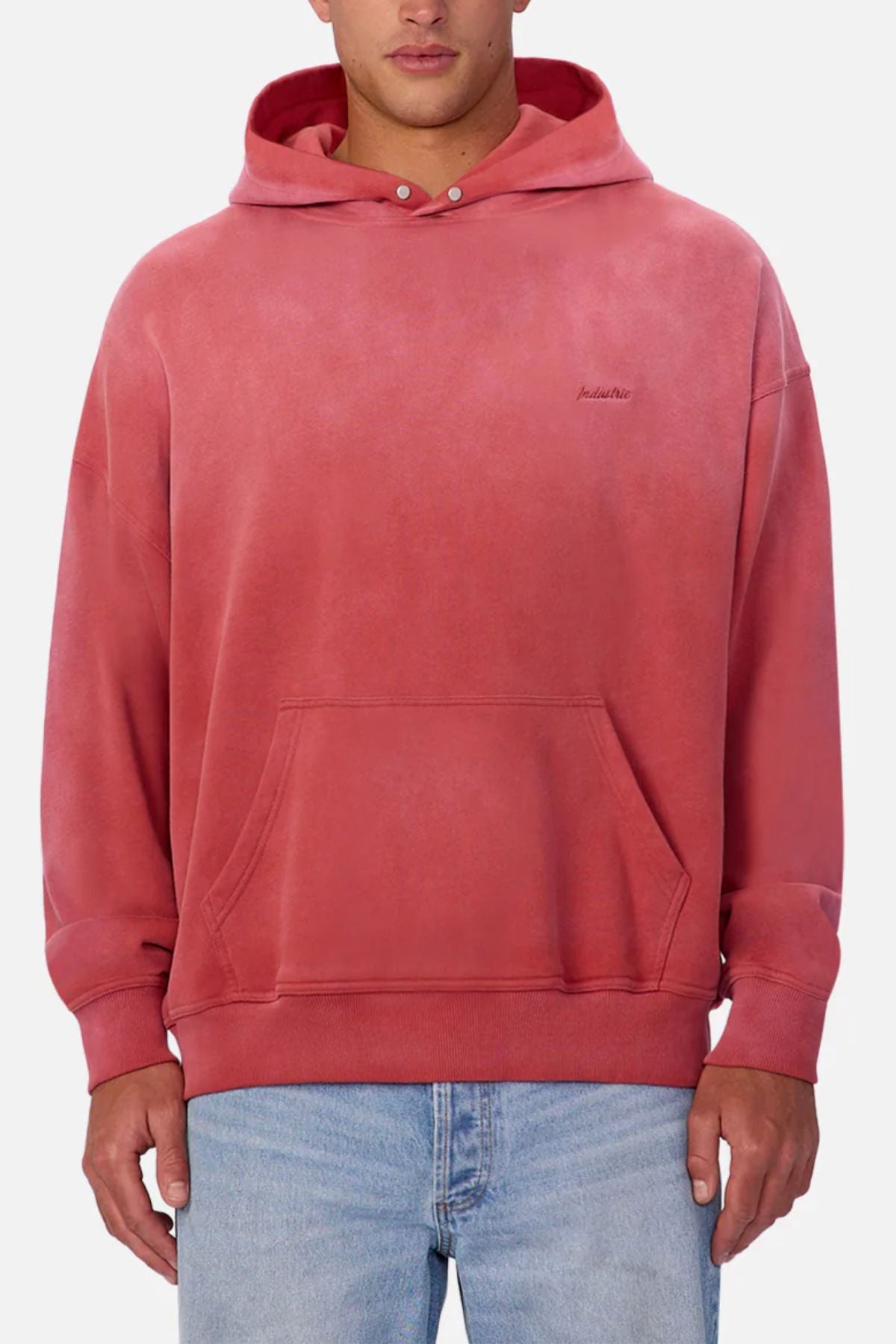 A man wearing the Industrie The Sandblast Hoodie in faded red with a front pocket and subtle left chest text, paired with blue jeans, stands against a plain light background. The 100% cotton hoodie is ideal for a laid-back look.