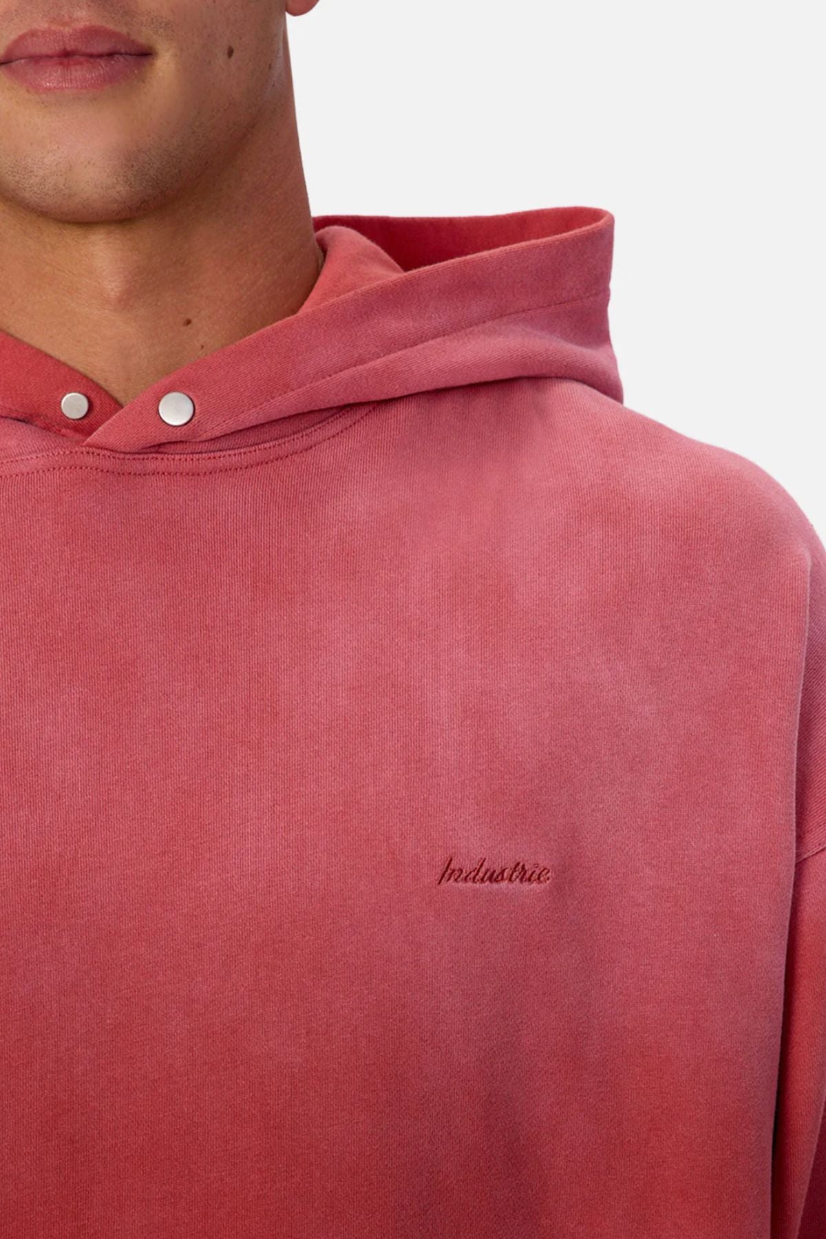 Someone wears the Industrie The Sandblast Hoodie in faded red, featuring silver snap buttons and "Industrie" embroidery on the chest. Made from 100% cotton, only their lower face and upper torso are visible, highlighting its relaxed style.