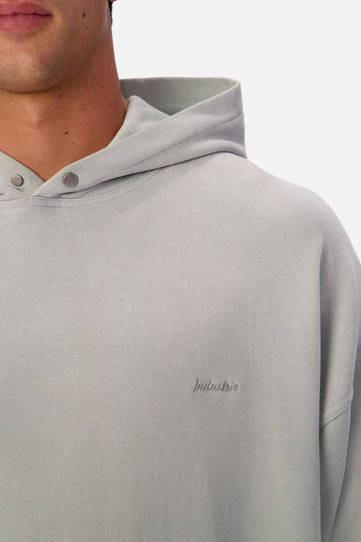 A person wears the Industrie The Sandblast Hoodie in light grey. It features a hood, snap buttons, and subtle "Industrie" chest embroidery. Made from 100% cotton, only the lower face and upper torso are visible for an easygoing look.