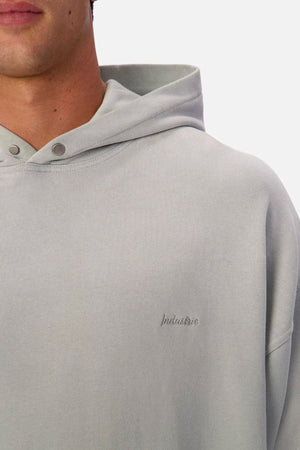 A person wears the Industrie The Sandblast Hoodie in light grey. It features a hood, snap buttons, and subtle "Industrie" chest embroidery. Made from 100% cotton, only the lower face and upper torso are visible for an easygoing look.