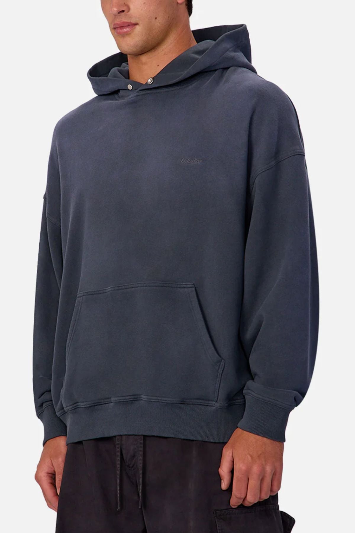 Wearing the Industrie The Sandblast Hoodie—a dark gray, 100% cotton hoodie with a front pocket and drawstring hood—paired with dark pants, this person stands against a plain light background in a laid-back style.