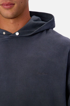 A close-up of a person wearing the Industrie The Sandblast Hoodie in dark blue cotton, featuring silver hood snap buttons and subtle “Produzione” embroidery on the chest. Set against a plain white background for a relaxed style.