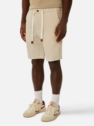 A person is standing wearing light beige, knee-length shorts with a drawstring waist, specifically the Industrie The Baller Linen Short featuring a single button at the waist. They also have on a beige t-shirt, white socks, and white sneakers with red accents. The background is plain white.