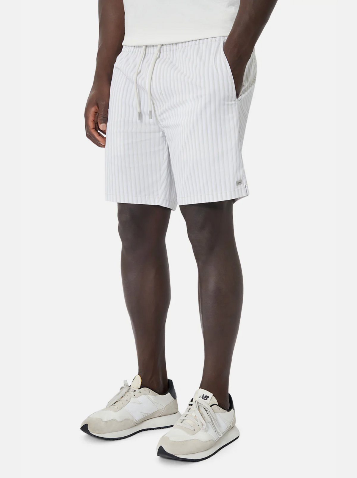 Wearing Industrie The Marchena Bahama Short with an elastic waist, a white shirt, and white athletic shoes with black soles, the person leans casually against a simple backdrop, perfectly capturing the relaxed summer vibe.