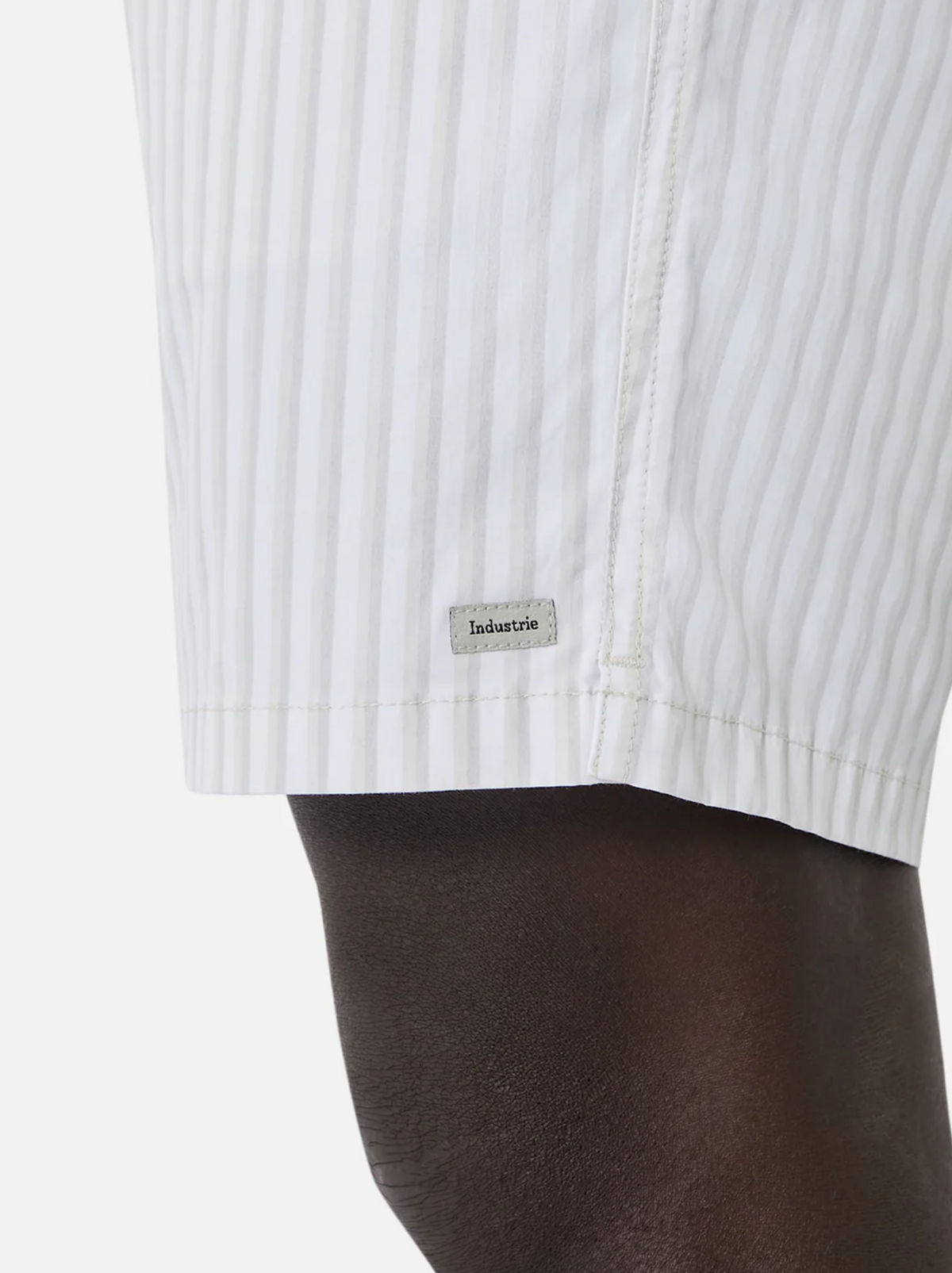 Close-up of a person wearing Industrie The Marchena Bahama Short, featuring white fabric with vertical light gray stripes and an elastic waist for comfort. A small rectangular "Industrie" tag is on the lower side, perfect for summer days against a plain white background.