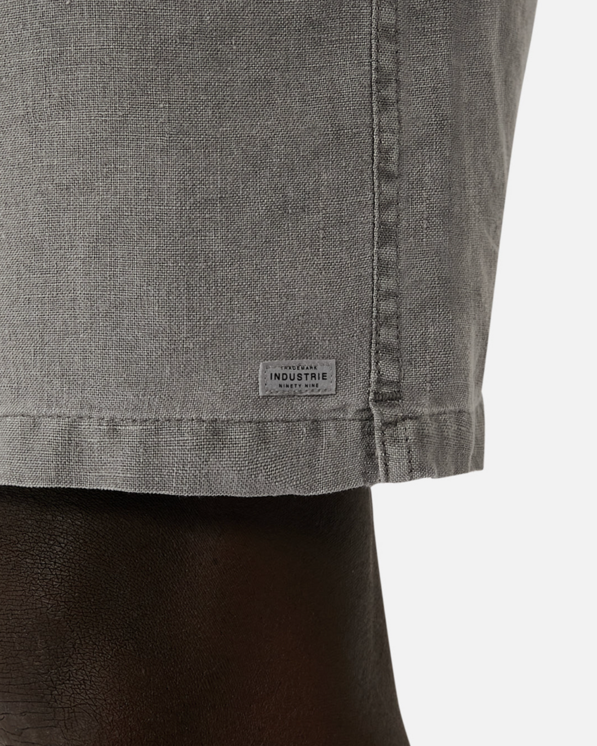 Close-up of gray fabric from the Industrie The Marina Linen Short, featuring a small rectangular "INDUSTRIE" label near the bottom edge. The 100% linen material indicates they are relaxed-fit shorts.