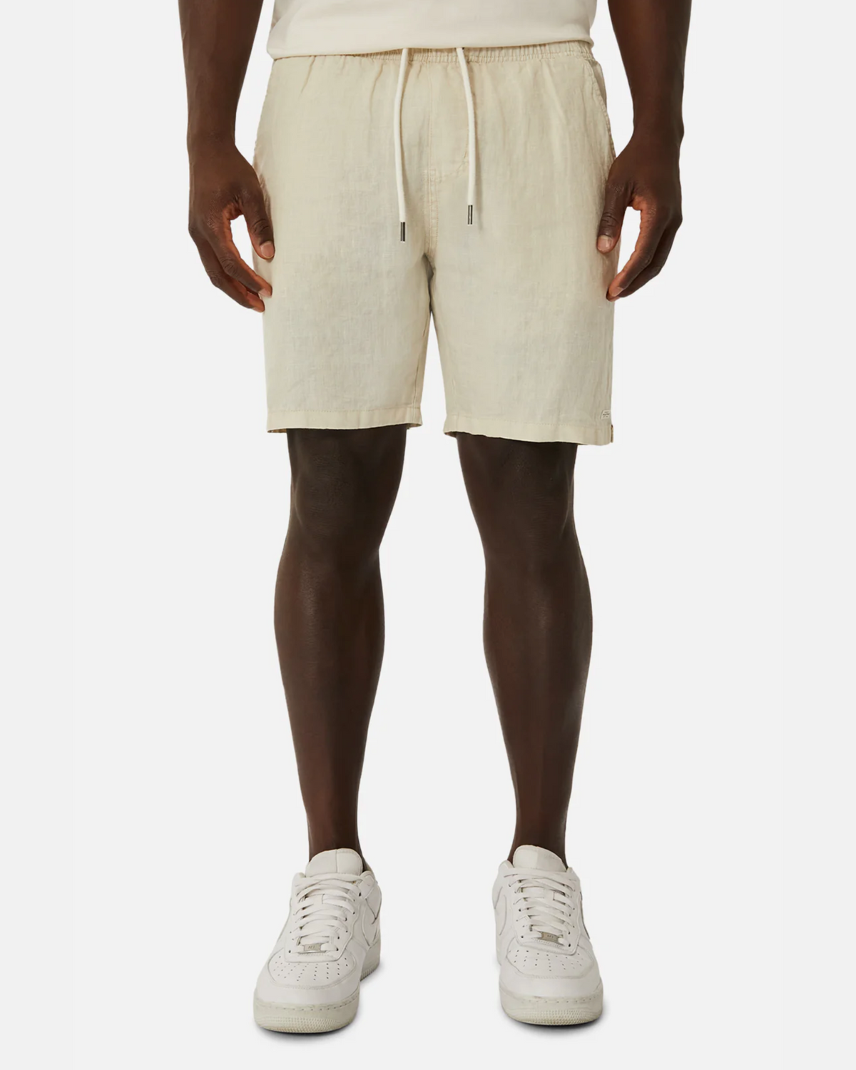 A person donning light beige Industrie The Marina Linen Shorts, crafted from pure linen with a drawcord waist, stands against a plain white background. Their hands are at their sides, revealing the waistband's drawstrings, perfectly paired with crisp white sneakers.