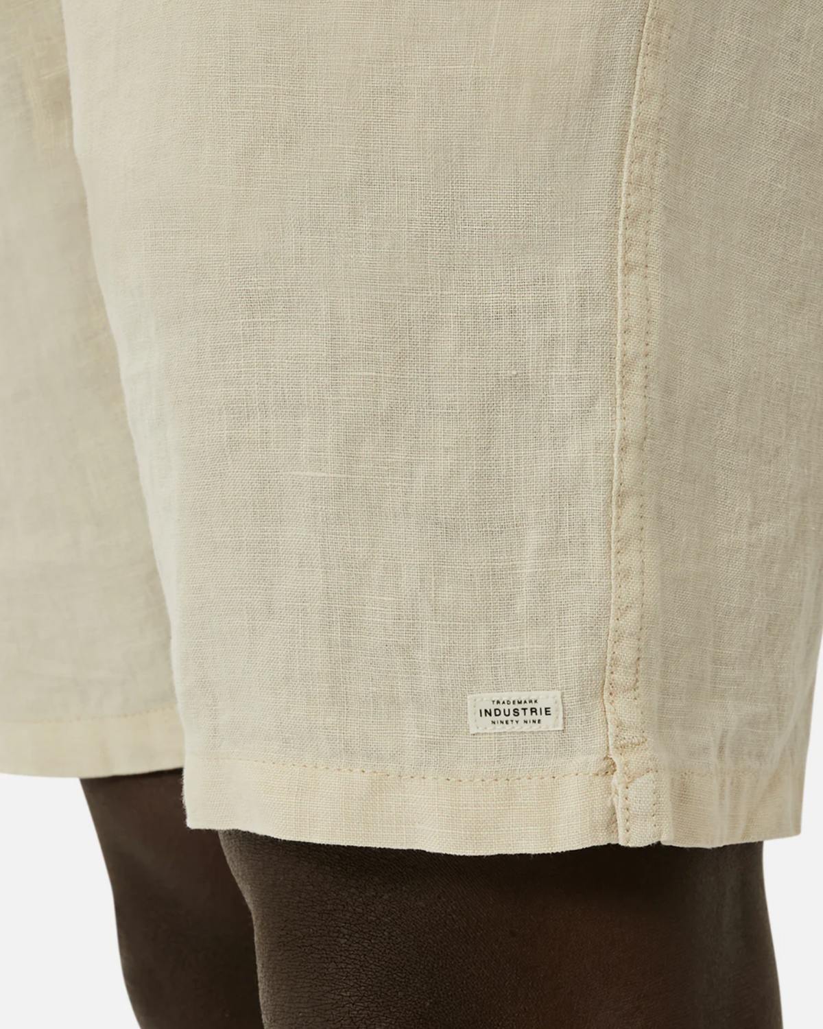 The close-up features the Industrie The Marina Linen Short made from pure linen with a drawcord waist. The beige shorts display a small white "INDUSTRIE" label on the hem against a plain white background.