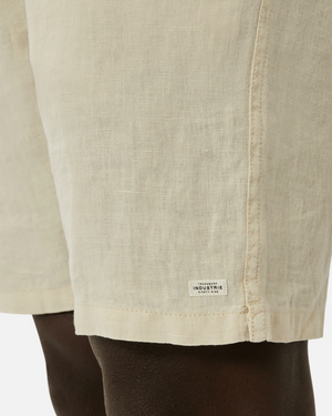 The close-up features the Industrie The Marina Linen Short made from pure linen with a drawcord waist. The beige shorts display a small white "INDUSTRIE" label on the hem against a plain white background.