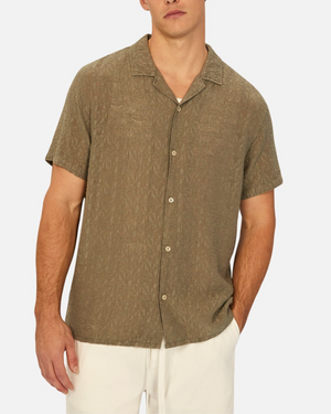 Someone in an Industrie Chiara Short Sleeve Shirt, olive green with a spread collar, stands against a white backdrop wearing white pants. The shirt's relaxed fit is ideal for casual outings, hands casually in pockets, exuding effortless style.