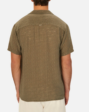 A person is shown from behind, wearing an olive green Industrie The Chiara Short Sleeve Shirt with a textured pattern. The shirt’s spread collar complements its loose fit, effortlessly paired with light-colored shorts against a simple white background.