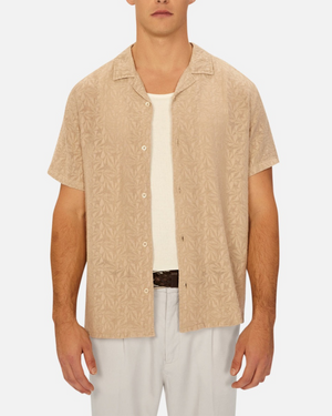 A man stands against a plain background wearing the Industrie The Chiara Short Sleeve Shirt in beige with an all-over pattern over a white undershirt, paired with light gray pants and a brown belt.