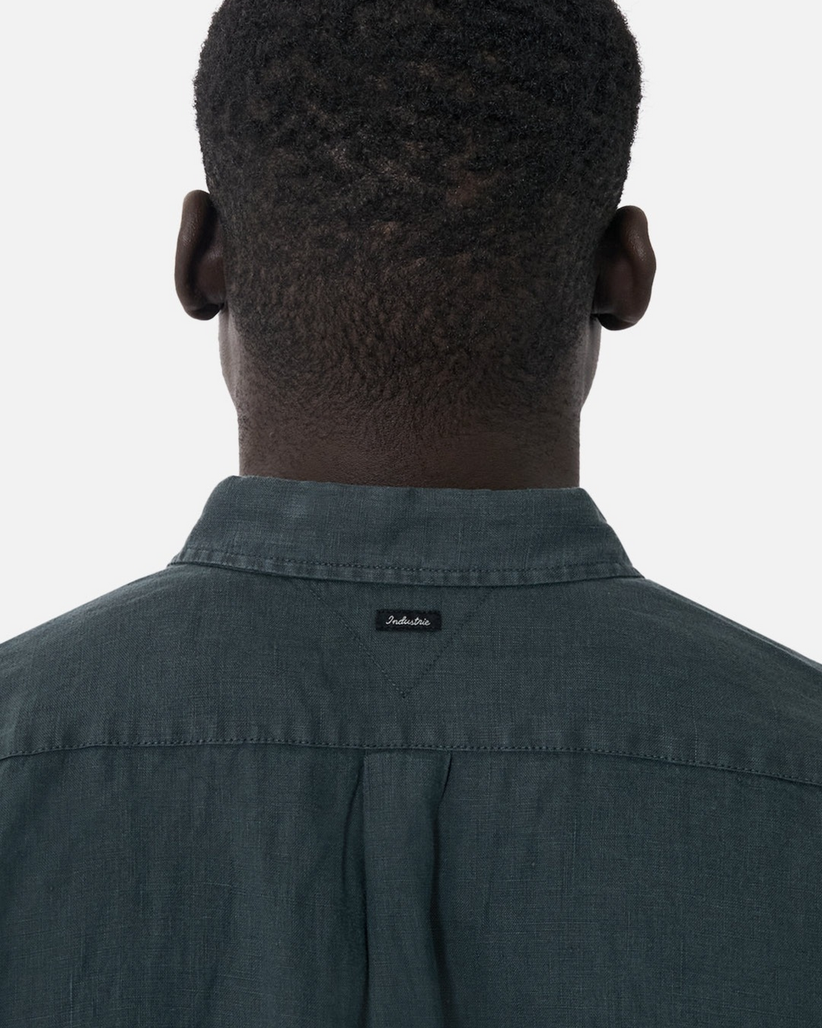 A person with short curly hair is seen from behind wearing the Industrie The Tennyson Linen Short Sleeve Shirt in dark green, featuring a small black label on the back collar. The background is plain white.