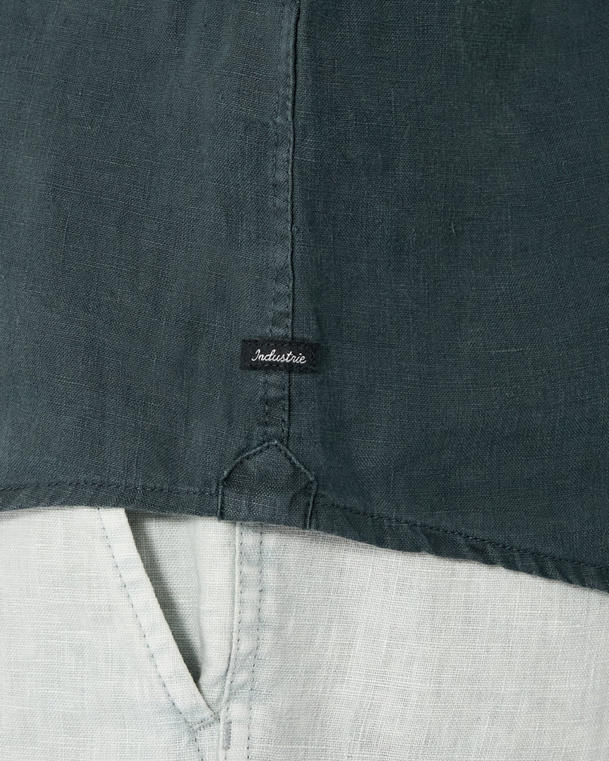 Close-up of the Industrie The Tennyson Linen Short Sleeve Shirt in dark green, featuring a center seam, short sleeves, and a small black "Industrie" tag above the hem, styled over light linen pants with a triangular stitch at the bottom edge.