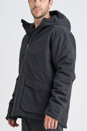 A man wears a black Jetpilot Onsite Pro Jacket with an adjustable drawstring hood and padded body, standing against a plain light background with his face partially turned away.