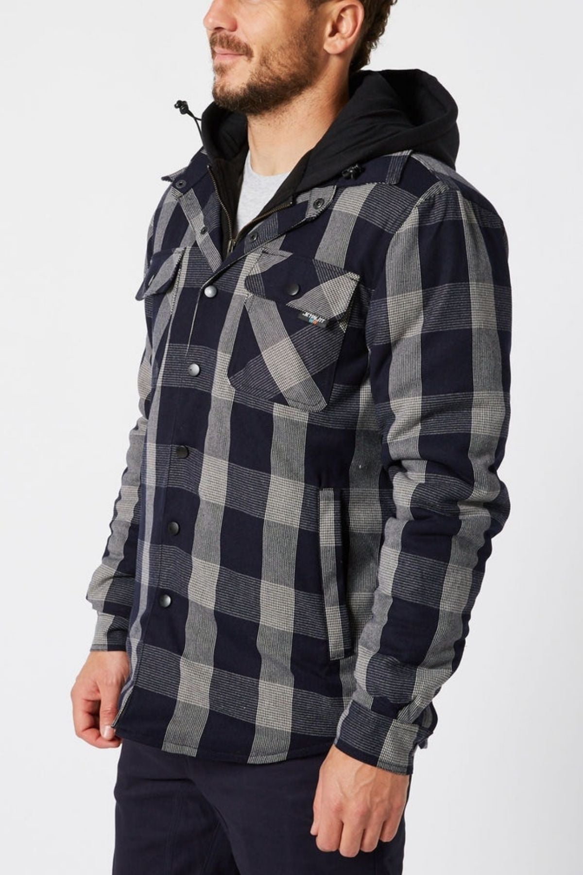 A man stands against a plain white background wearing a Jetpilot Quilted Flannel Jacket in black and gray plaid with a hood, buttoned front, and front pockets.