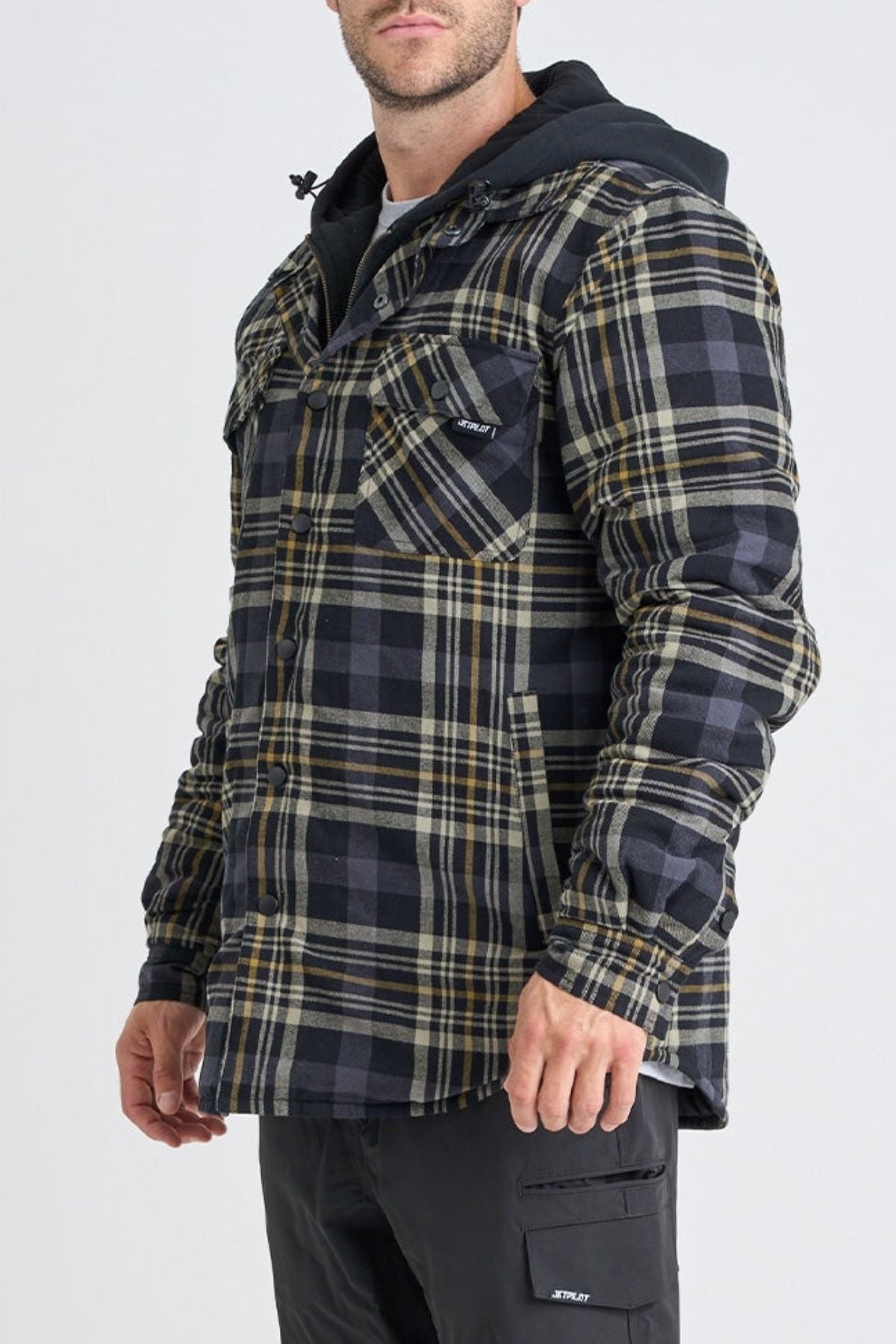 A man wearing a Jetpilot Quilted Flannel Jacket in black and yellow with a hood, buttoned chest pockets, and black pants stands against a plain light background.