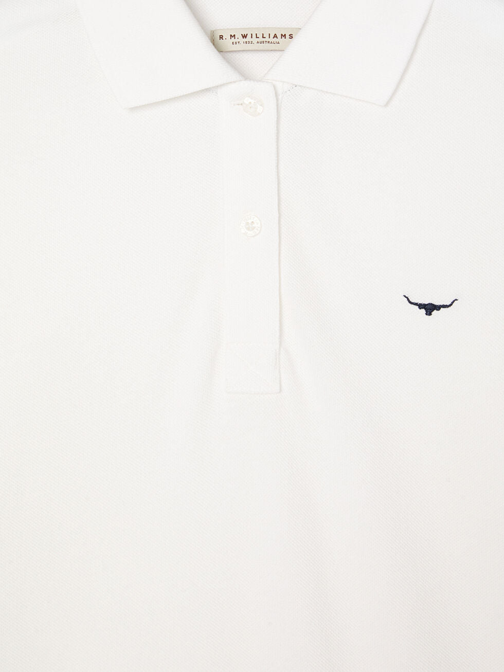 Close-up of the RM Williams Margaret Polo in white pique cotton, showing a small dark bull logo on the left chest, three white buttons on the placket, and an inner label with "R.M. WILLIAMS.