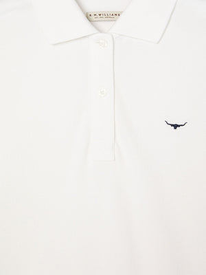 Close-up of the RM Williams Margaret Polo in white pique cotton, showing a small dark bull logo on the left chest, three white buttons on the placket, and an inner label with "R.M. WILLIAMS.