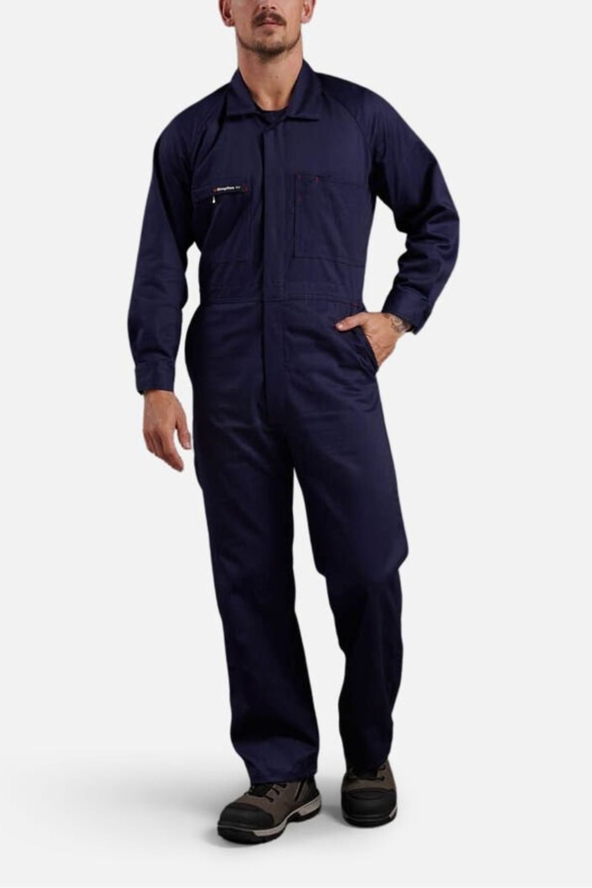 A man wearing King Gee Combination Drill Overall stands against a plain light background, with one hand in his pocket and dark work shoes.