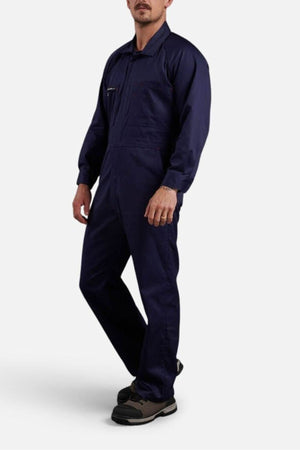 A person wears the King Gee Combination Drill Overall in durable navy blue cotton with a front zipper and chest pocket, paired with black-and-gray work shoes, standing against a plain light background, face partially visible.