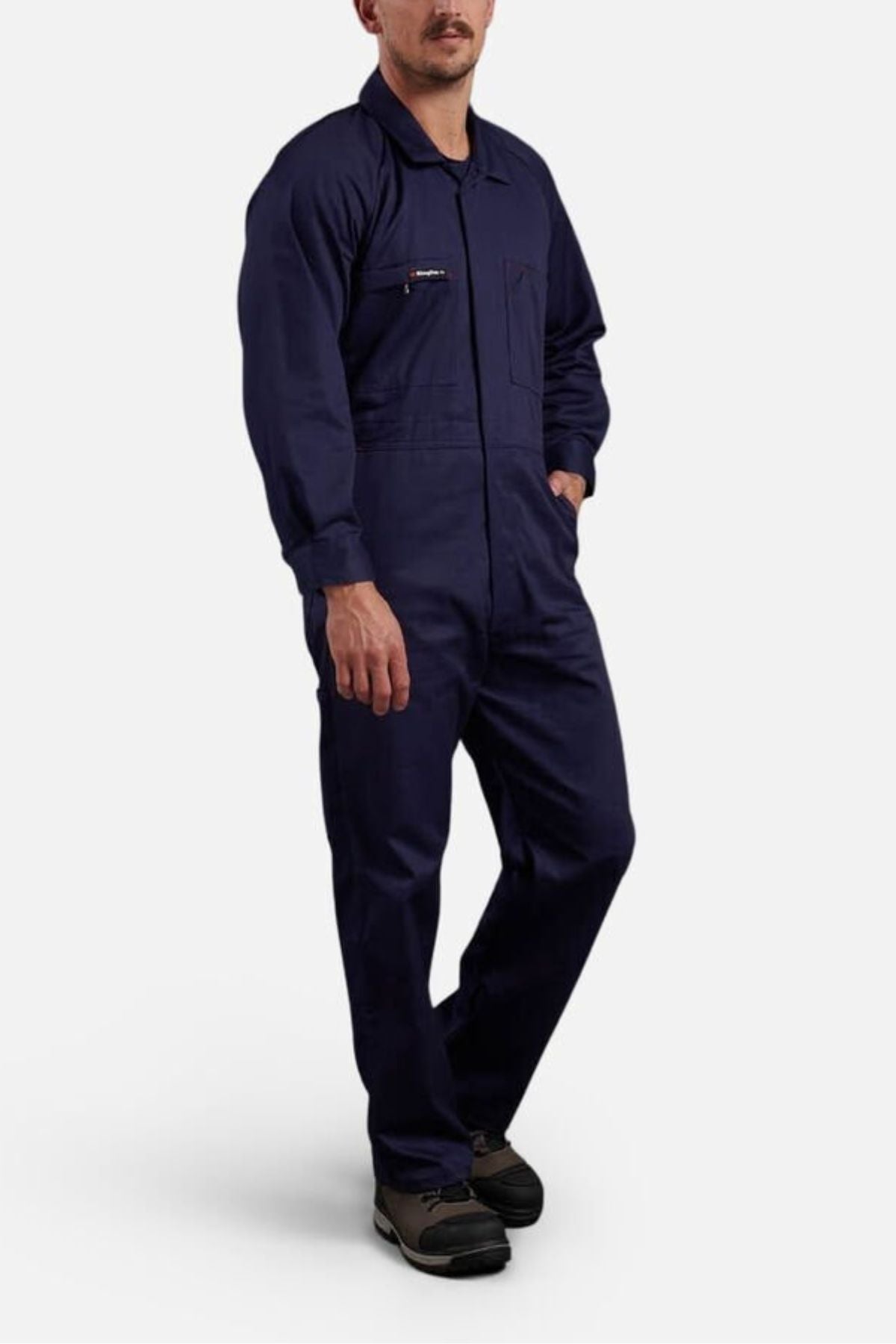A person wears King Gee Combination Drill Overalls, featuring a zippered chest pocket, with black shoes, standing with one hand in their pocket against a plain light background.