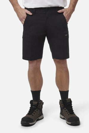 A person wearing black King Gee Workcool Pro Shorts, a white shirt, black socks, and brown hiking boots stands with hands in pockets against a plain white background.