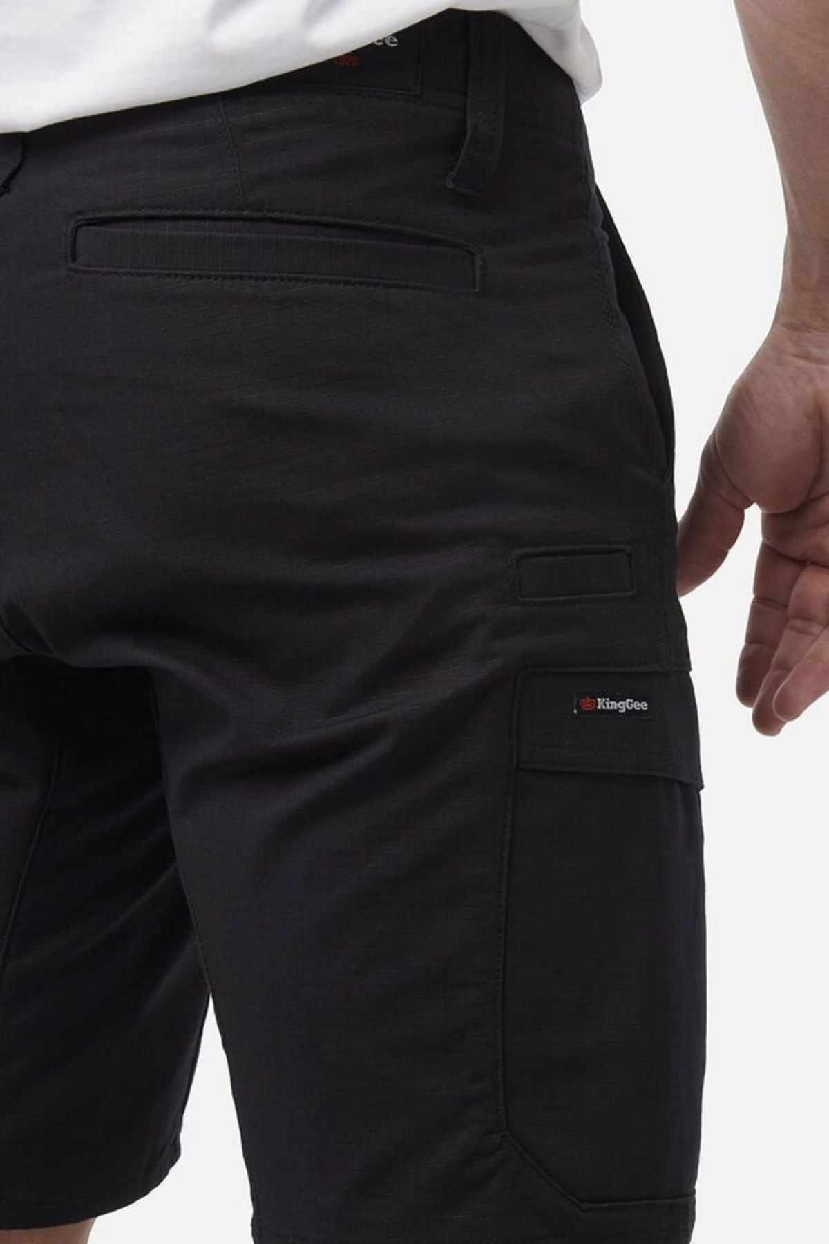 Someone wears black King Gee Workcool Pro Shorts made from ripstop stretch fabric, with multiple pockets—including a side pocket featuring a red and white KingGee logo tag. Their right arm is relaxed at their side against a plain white background.