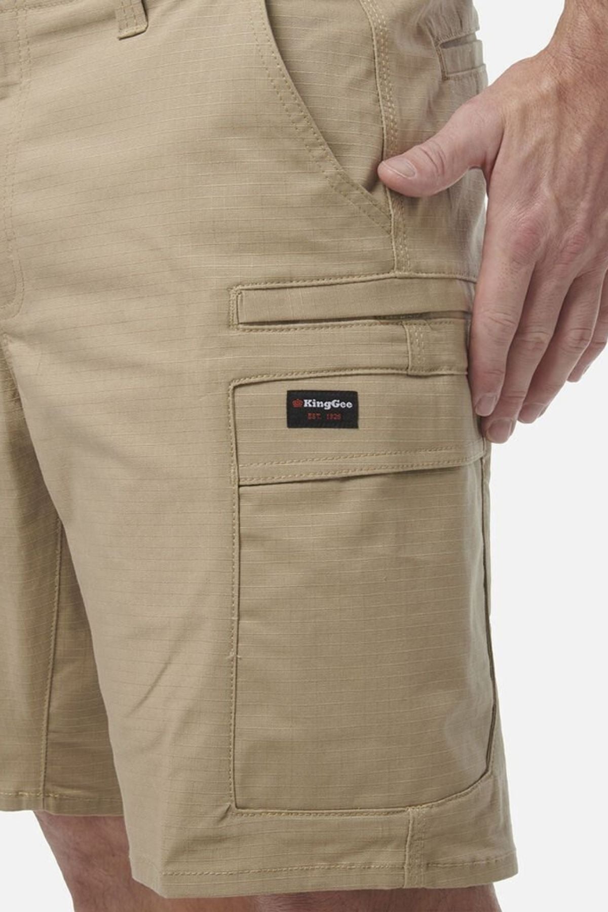 A close-up of a person wearing tan King Gee Workcool Pro Shorts made from ripstop stretch fabric, featuring side pockets and a KingGee logo patch above one pocket, with one hand resting by their side.