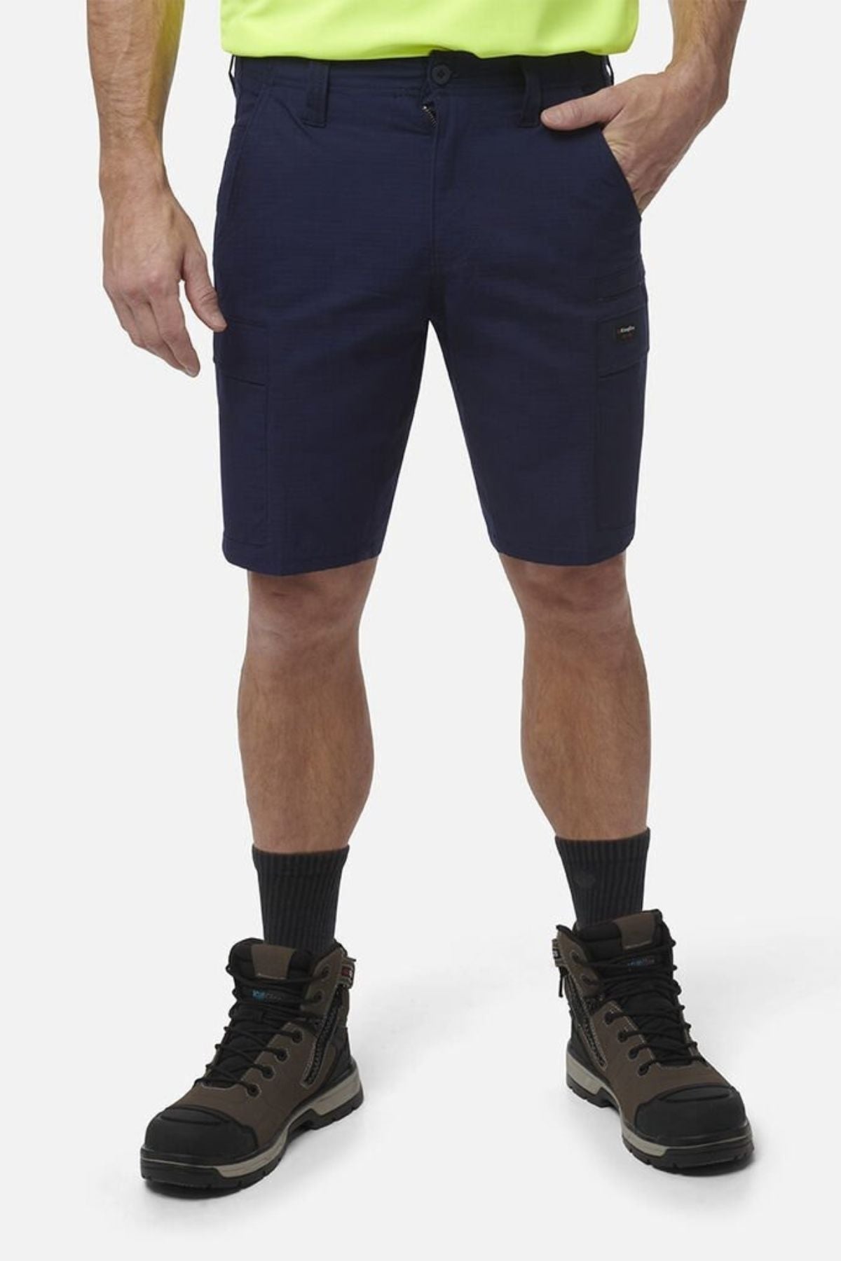 A person stands against a plain white background wearing King Gee Workcool Pro Shorts in navy, paired with a neon yellow shirt, black socks, and brown work boots. Only the lower half of their body is visible.