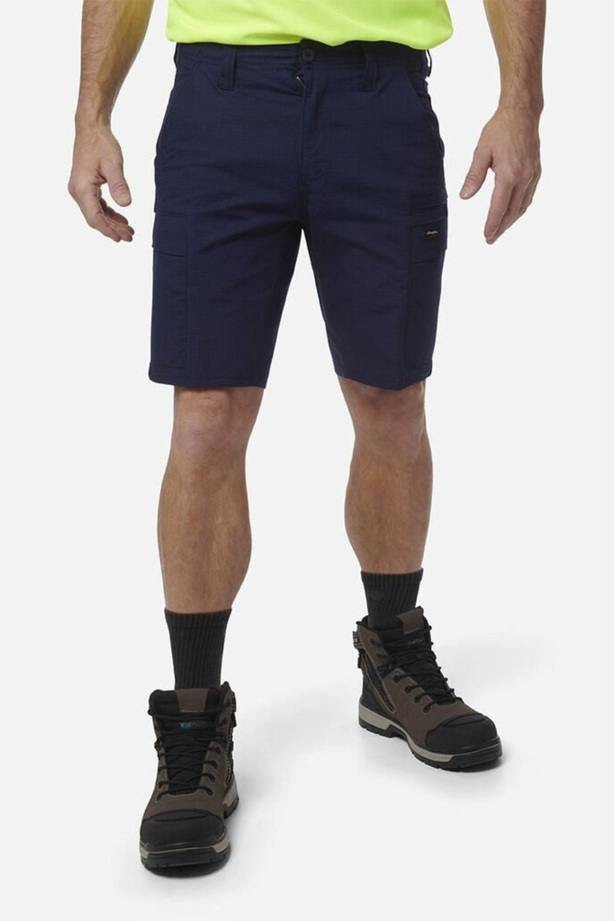 A person wearing navy blue King Gee Workcool Pro Shorts made from durable Ripstop stretch fabric, a neon yellow shirt, black socks, and brown work boots stands against a plain white background. The person's head is not visible in the image.