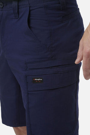 A person wears dark blue King Gee Workcool Pro Shorts, made from durable material with multiple pockets, including a cargo pocket featuring the KingGee logo tag on the right thigh. Their right hand is visible near the shorts.
