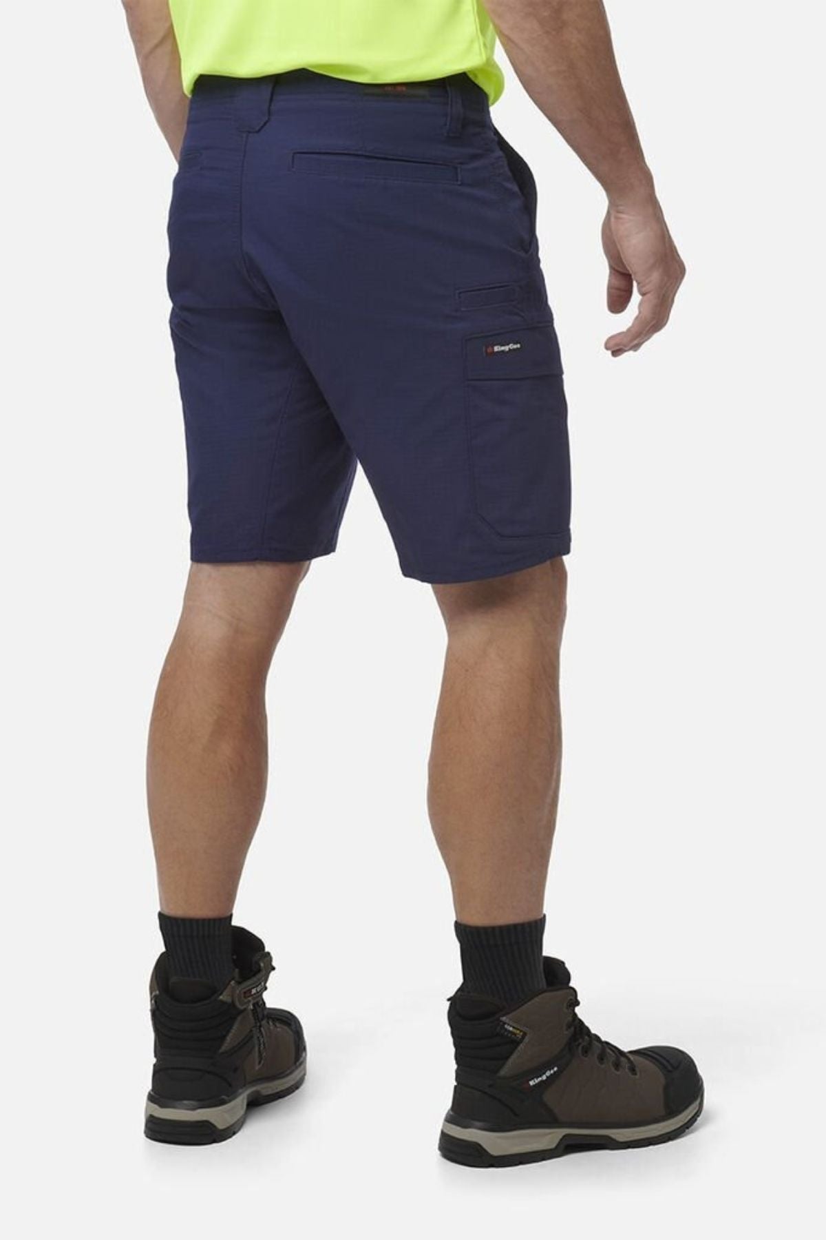 A person wearing King Gee Workcool Pro Shorts in navy blue, a neon yellow shirt, black socks, and brown work boots stands with their back to the camera against a plain white background.