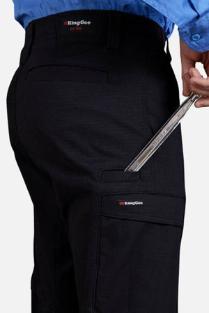 Wearing black King Gee Workcool Pro Stretch Cargo Pants and a blue shirt, a person places a metal wrench in a side pocket. The pants feature multifunctional pockets for convenience, with the King Gee logo clearly visible.