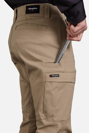 A person, visible from the waist down, places a folding ruler into the tool pocket of King Gee Workcool Pro Stretch Cargo Pants in tan.