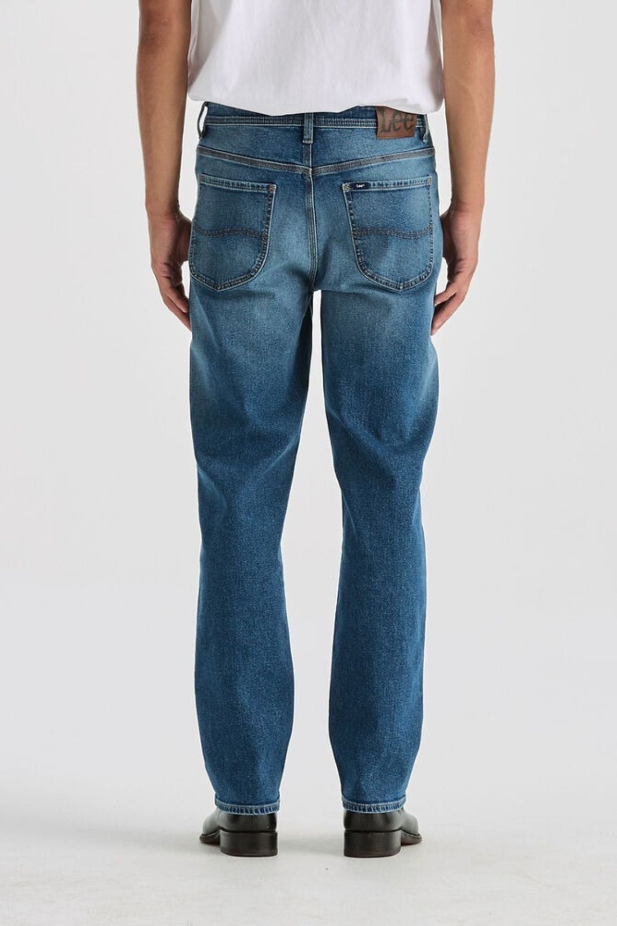 A person wearing Lee Originals L101 Straight Jean in blue and a white t-shirt stands facing away from the camera. They have on black shoes, against a plain light-colored background.
