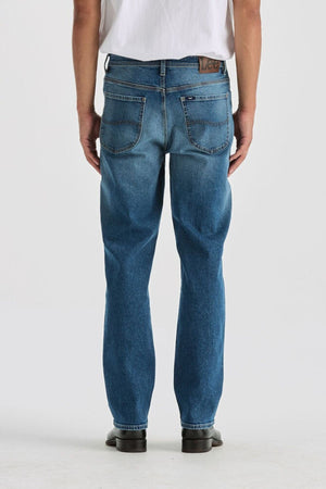 A person wearing Lee Originals L101 Straight Jean in blue and a white t-shirt stands facing away from the camera. They have on black shoes, against a plain light-colored background.