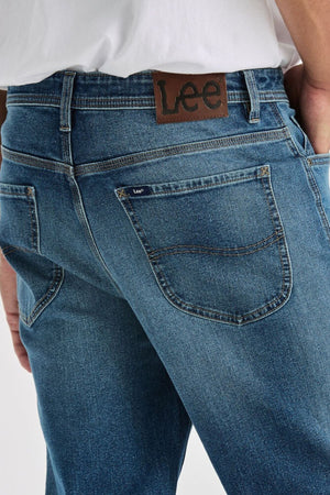 A close-up shows someone in a white shirt wearing blue Lee Originals L101 Straight Jeans. The leather Lee patch appears above the right back pocket, and their hand is partially tucked into the pocket.
