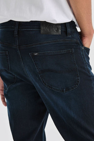 Close-up of a person wearing Lee Originals L100 Slim Jean in dark blue with a white shirt, hand in back pocket. The slim design ensures comfort and features a black leather "Lee" patch above the right pocket.