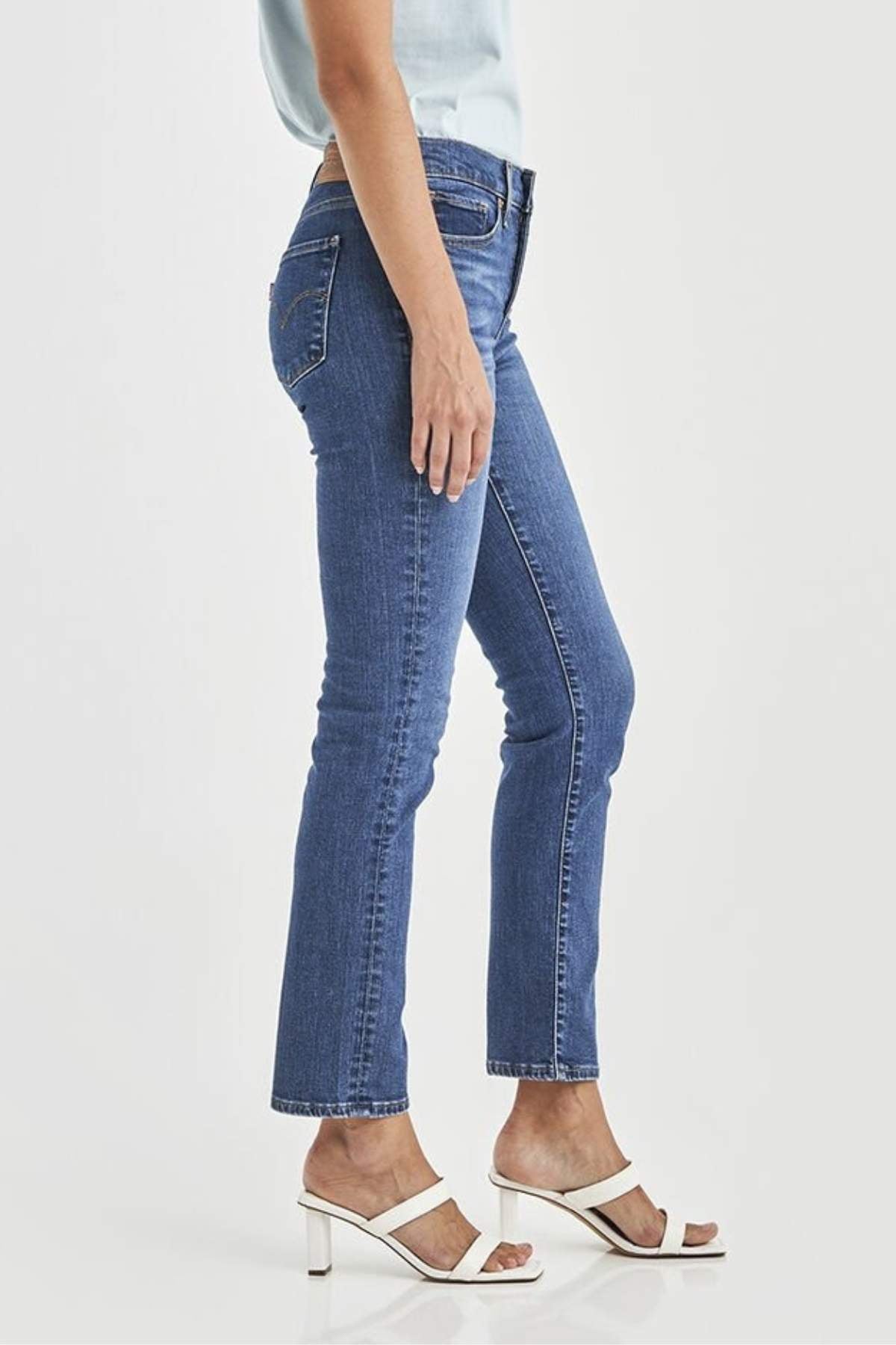 Wearing Levis 312 Shaping Slim Jean, a light blue top, and white heeled sandals, a person stands side-on against a plain white background with only the lower half of their body visible.