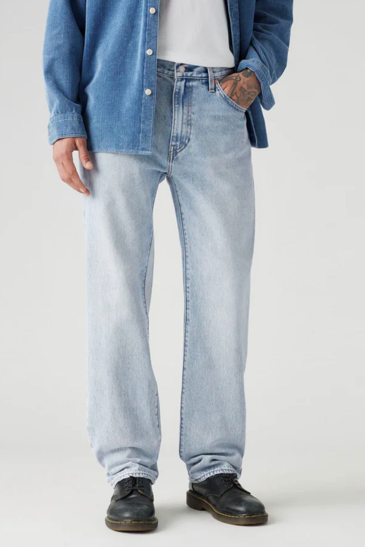 Wearing Levis 555 Relaxed Straight Jean in light blue non-stretch denim, paired with a white T-shirt, blue button-up shirt, black shoes, and displaying a tattoo on their left hand, the person stands confidently.