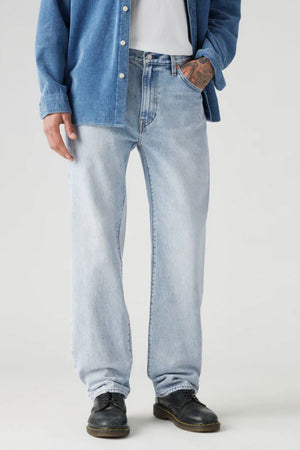 Wearing Levis 555 Relaxed Straight Jean in light blue non-stretch denim, paired with a white T-shirt, blue button-up shirt, black shoes, and displaying a tattoo on their left hand, the person stands confidently.