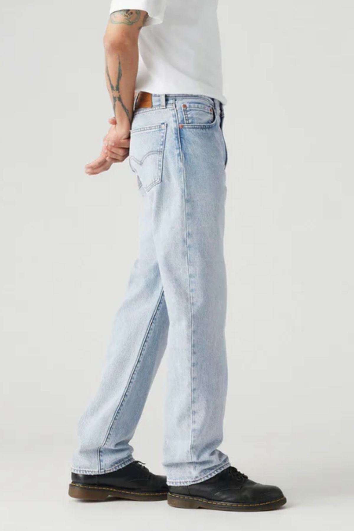 A person stands sideways in Levis 555 Relaxed Straight Jeans in light blue, paired with a white shirt and black shoes. Their tattooed forearm is visible, one hand behind their back, against a plain light-colored background.