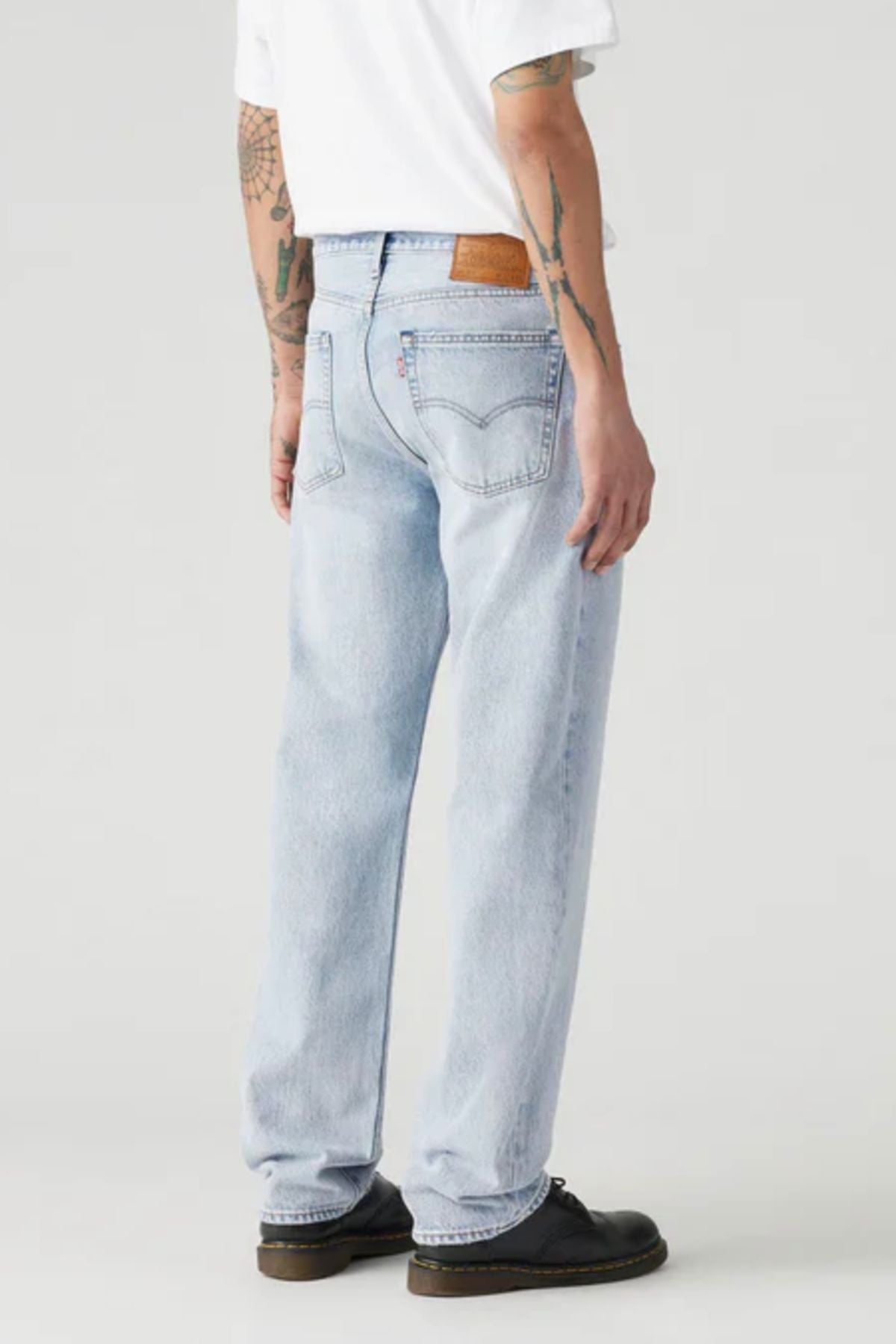 Someone stands facing away in Levis 555 Relaxed Straight Jean (light blue) and a white T-shirt, showing tattooed arms and black shoes. The background is plain and light-colored.