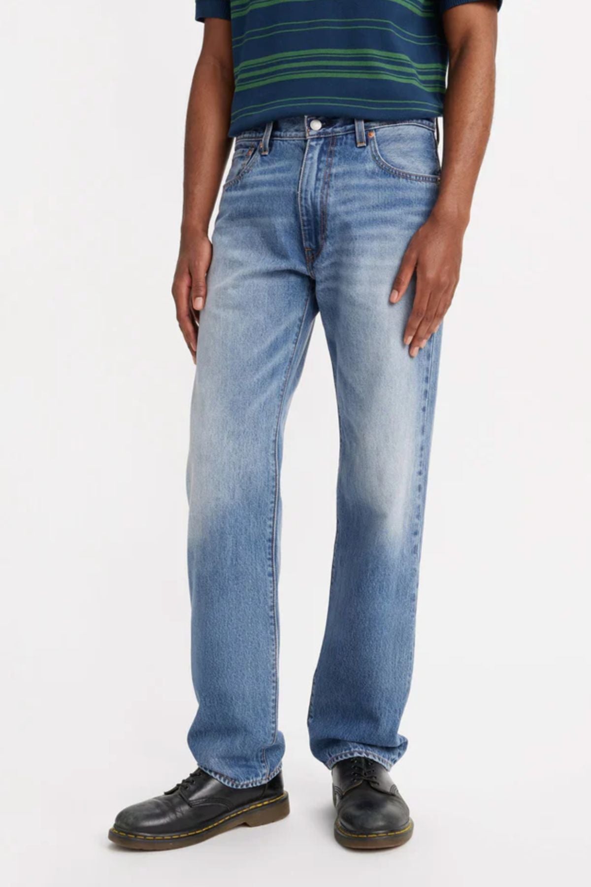 A person stands against a plain light background, shown from shoulders down, wearing light blue Levis 555 Relaxed Straight Jeans, a dark belt, black shoes, and a dark striped short-sleeve shirt.