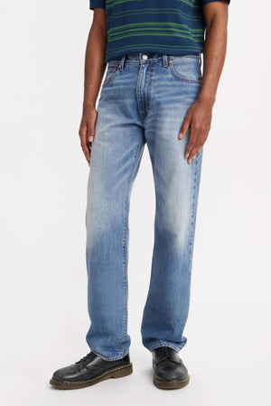 A person stands against a plain light background, shown from shoulders down, wearing light blue Levis 555 Relaxed Straight Jeans, a dark belt, black shoes, and a dark striped short-sleeve shirt.