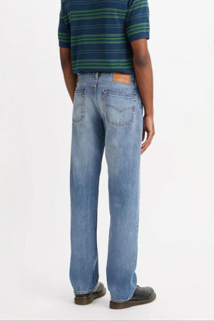A person faces away wearing a green and navy striped short-sleeve shirt, Levis 555 Relaxed Straight Jean in light blue denim, and dark shoes against a plain white background.