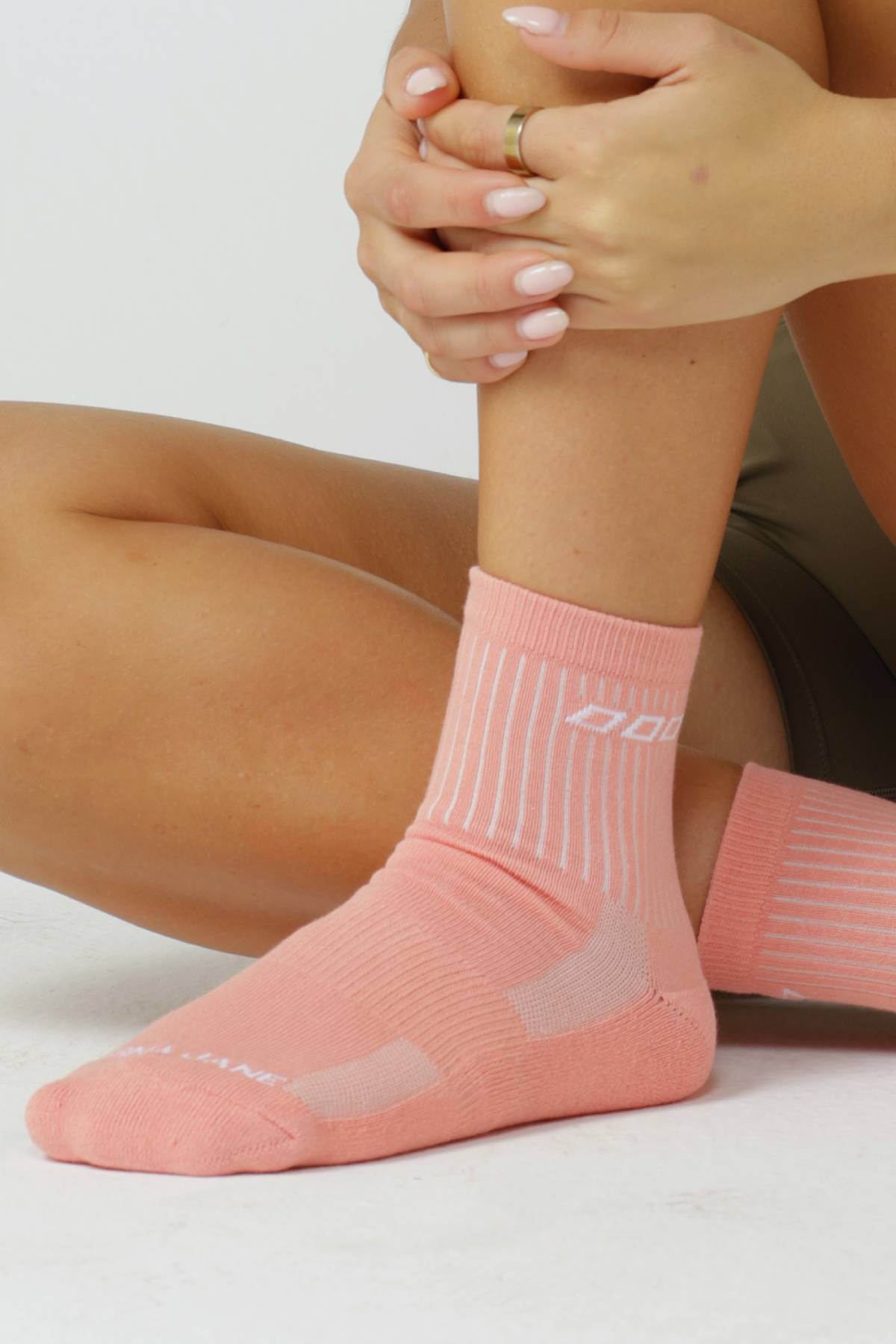 A person wears Lorna Jane 3pk LJ Quarter Crew Socks in coral pink, made from a breathable cotton blend with white stripes and text, sits with one knee up and light pink fingernails clasped around their shin.