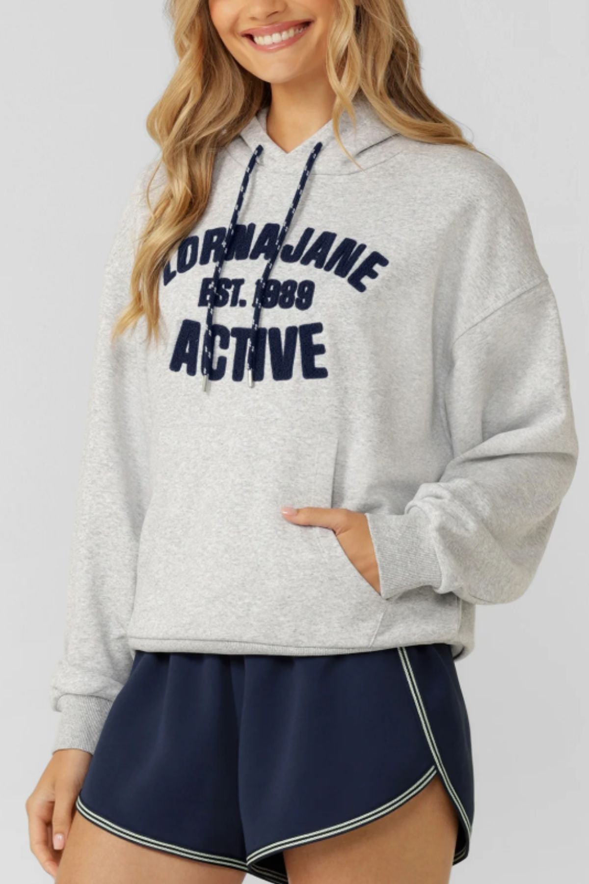 A smiling woman wears a vintage-inspired Lorna Jane Active Club Hoodie featuring "LORNA JANE EST. 1989 ACTIVE" in navy, paired with matching navy athletic shorts with white trim.